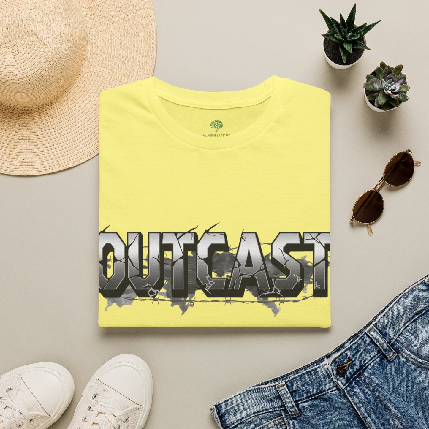 A folded pale yellow T-shirt with a large metallic-style 'OUTCAST' graphic across the chest